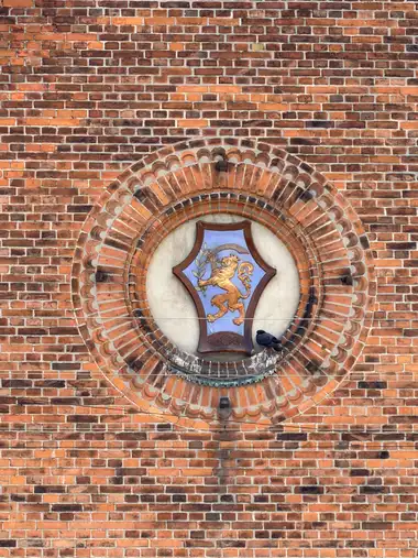 A painted heraldic shield with a rearing golden lion holding a leafy branch is set within a circular niche of concentric moulded brickwork.