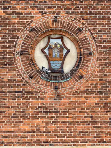 A painted heraldic shield is set within a circular niche of concentric moulded brickwork on the red-brick facade.