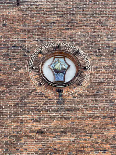 A painted shield depicting a sailing ship above blue waves is set in a circular niche framed by concentric courses of moulded red brick.