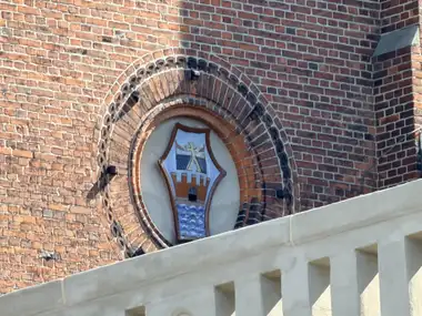 A painted heraldic shield with a crenellated gateway and waves is set in a circular recess framed by concentric courses of moulded red brick.