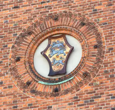 A painted heraldic shield with three blue lions and red hearts is mounted in a circular recess framed by concentric courses of moulded red brick.