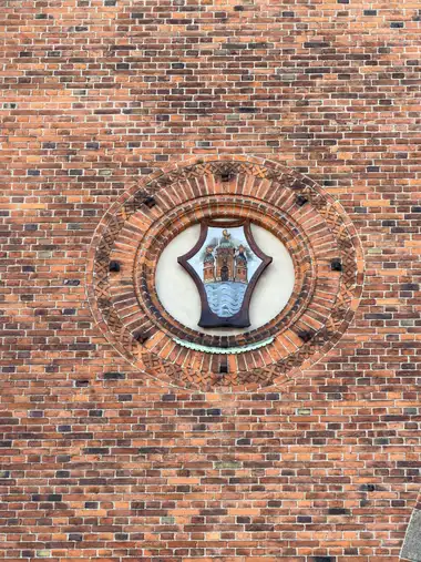 A painted Copenhagen coat of arms is set in a circular recess framed by concentric moulded and floral red-brick courses within a broad brick wall.