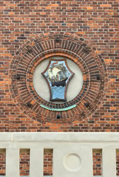 A painted shield bearing a sailing ship is set within a deep circular niche of concentric moulded brick courses and carved terracotta ornament.