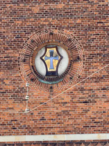 A painted heraldic shield with a pale blue cross on a gold field is set in a circular recess framed by concentric courses of moulded red brick.