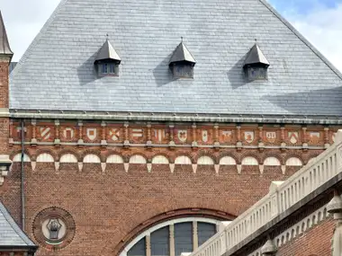 A long frieze of small geometric shield panels runs beneath the eaves of a red-brick wall below a steep slate roof with three dormers.