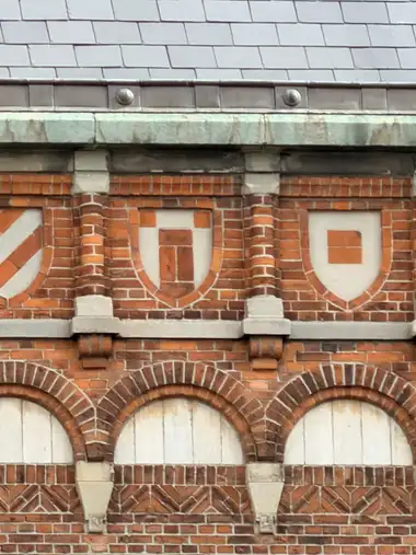 A close view of repeating red-tile shield panels in white plaster between moulded brick piers, beneath a pale stone cornice and slate roof.
