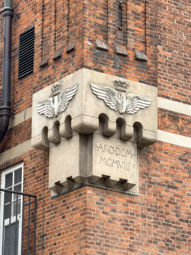 A sandstone corner console cut with ·AÑO·DOM· MCMVIII, beneath a band of crowned winged wheels.