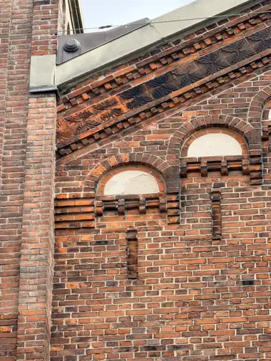 A close view of a red-brick gable with corbelled courses, two blind arched recesses, and a diagonal band of dark vegetal relief tiles beneath the coping.