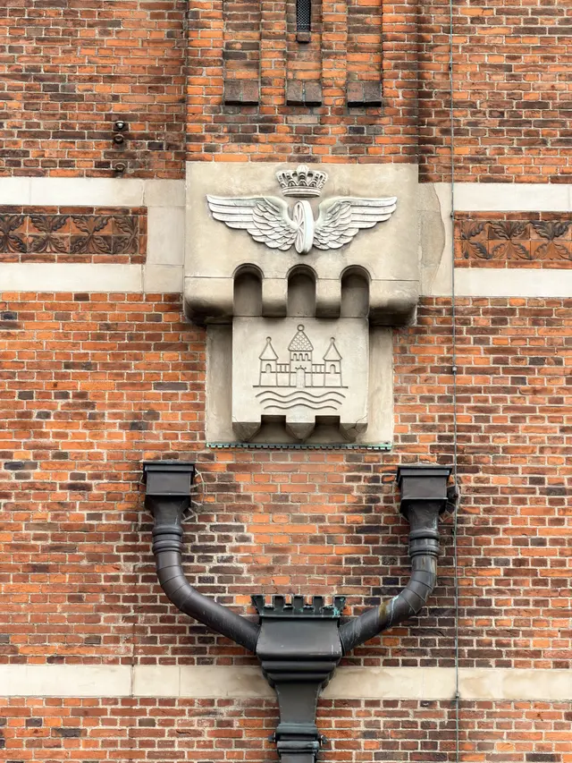A winged wheel under a crown carved in pale stone, set above a panel showing three towers over water.