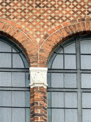 A close view shows paired arched windows divided by a brick pier, with radiating brick voussoirs and a repeating red-brick floral diaper pattern set against pale plaster above.