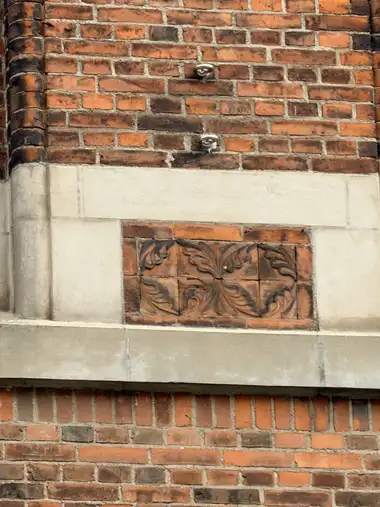 A rectangular panel of moulded terracotta tiles with stylised foliage is set within pale stone banding and surrounding red brickwork.