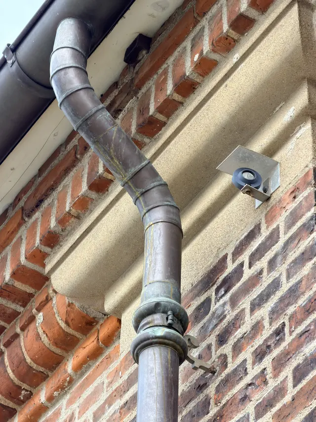 A metal rainwater downpipe curving out and back around a pale stone band, with a thickened moulded collar at the joint.