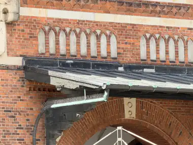A red-brick wall with two bands of pointed blind arches, a leaf-patterned terracotta frieze, pale stone banding, and the upper part of an entrance canopy.
