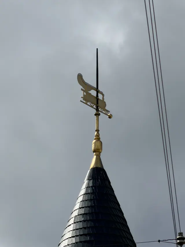 A gilded weather vane in the shape of a steam locomotive, with spoked wheels and a curling plume of smoke, on top of a slate spire.