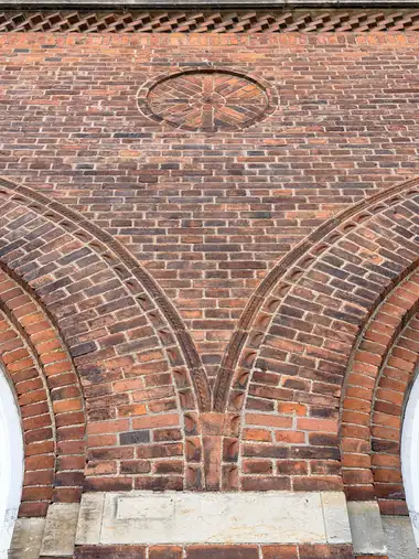 A close frontal view shows richly patterned red-brick masonry with intersecting moulded arches, a circular radial brick medallion, and a perforated brick cornice.