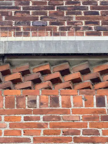 Close view of red-brick masonry with a projecting angled brick corbel course beneath a concrete band fitted with bird-deterrent spikes.
