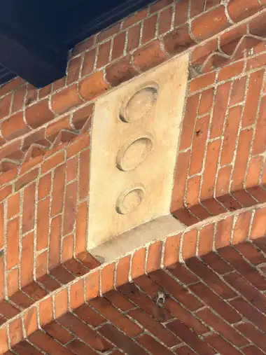 Decorative red brickwork surrounds a pale rectangular stone panel bearing three projecting circular bosses beneath a dark overhang.