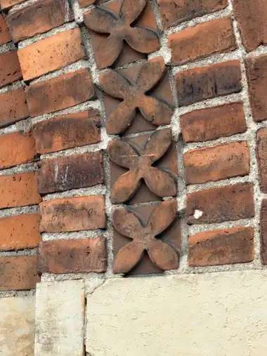 A vertical band of projecting four-lobed leaf-shaped terracotta ornaments is set into weathered red-brick masonry above a pale rendered base.