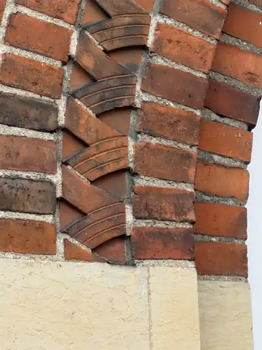 A close view of a vertical band of diagonally laid moulded bricks within weathered red-brick masonry above a pale stone base.