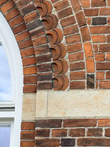 A close view shows irregular red brickwork beside a window arch, with a vertical row of semicircular moulded bricks and pale stone banding below.