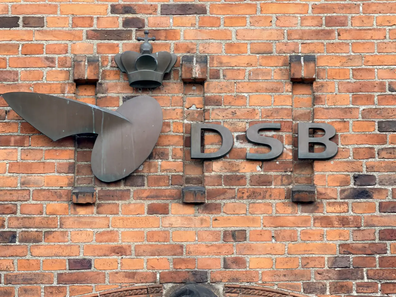 A wrought-iron DSB sign with a winged wheel, fixed to red brick.