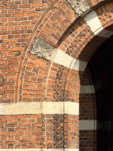 A close view shows a red-brick entrance arch with moulded floral brick bands, pale stone courses and carved floral stone panels.