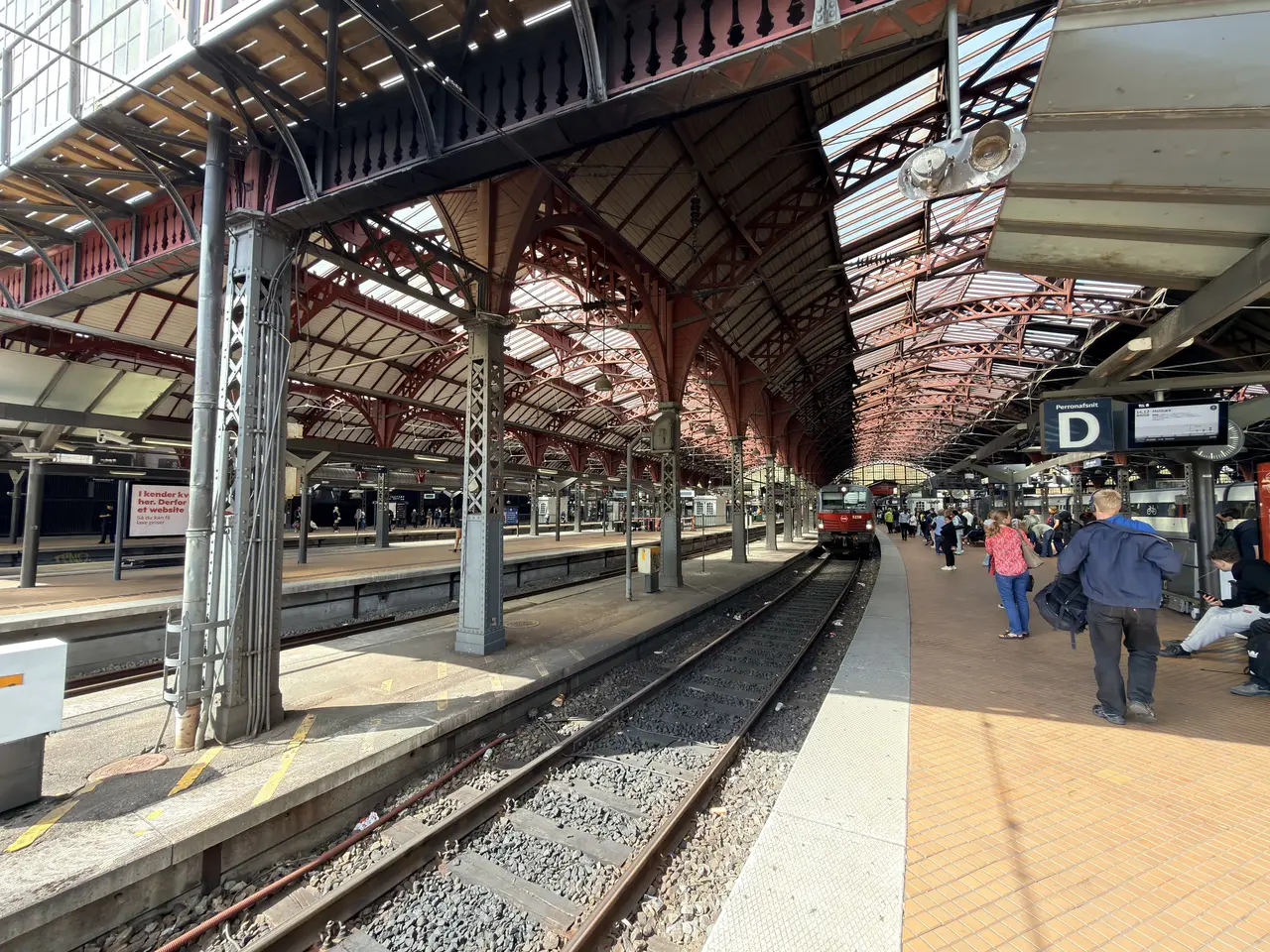 The platform under the timber arches of the platform hall, with a train, travellers and a sign gantry.