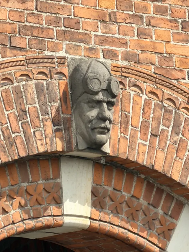A carved man's head in a leather helmet with round goggles pushed up on the forehead, set as a keystone over a brick arch.