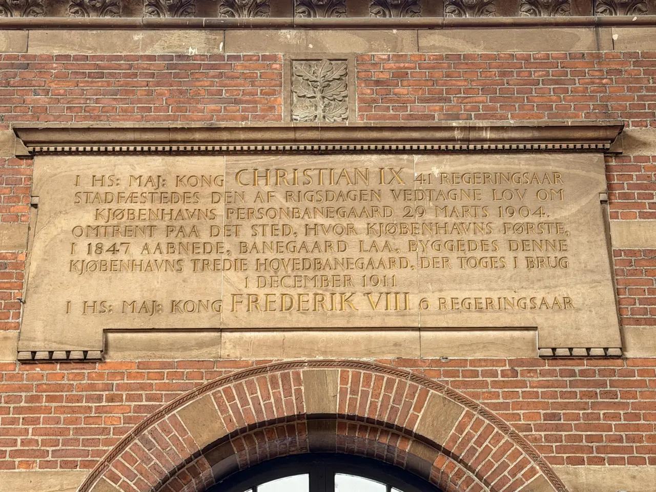 A large sandstone inscription tablet with gilded capitals, set in the brickwork above a round-arched opening in the main hall.