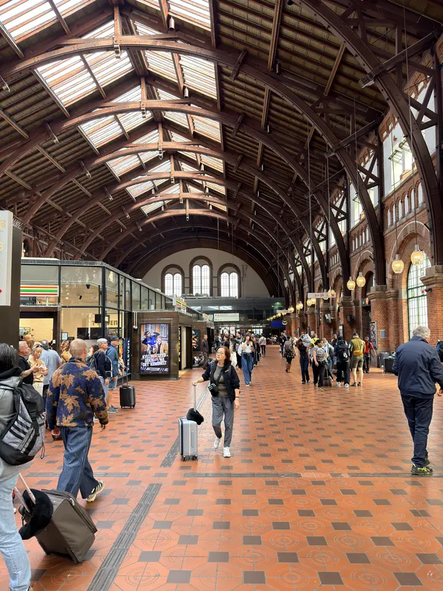 The main hall seen from the floor: the timber arched roof with long rooflights, brick side walls with round-arched windows, glazed shop units built in along one side, a patterned tiled floor, and travellers crossing it.