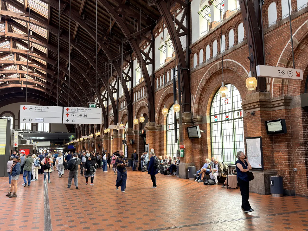 The main hall: timber arches over brick arcaded walls, hanging lamps, signs and travellers on the tiled floor.