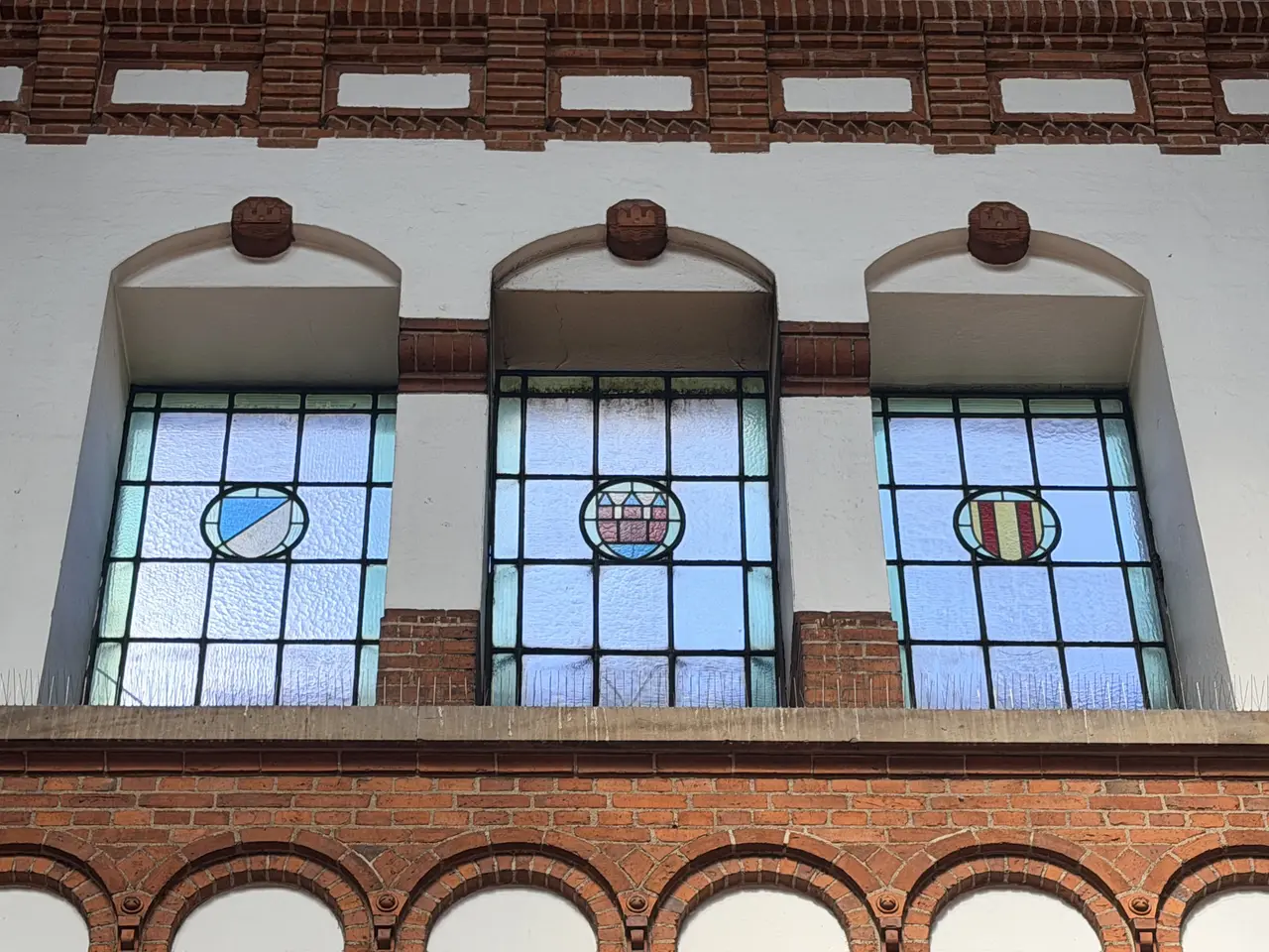 Three arched windows high in the great hall, their leaded glazing carrying small painted heraldic shields.