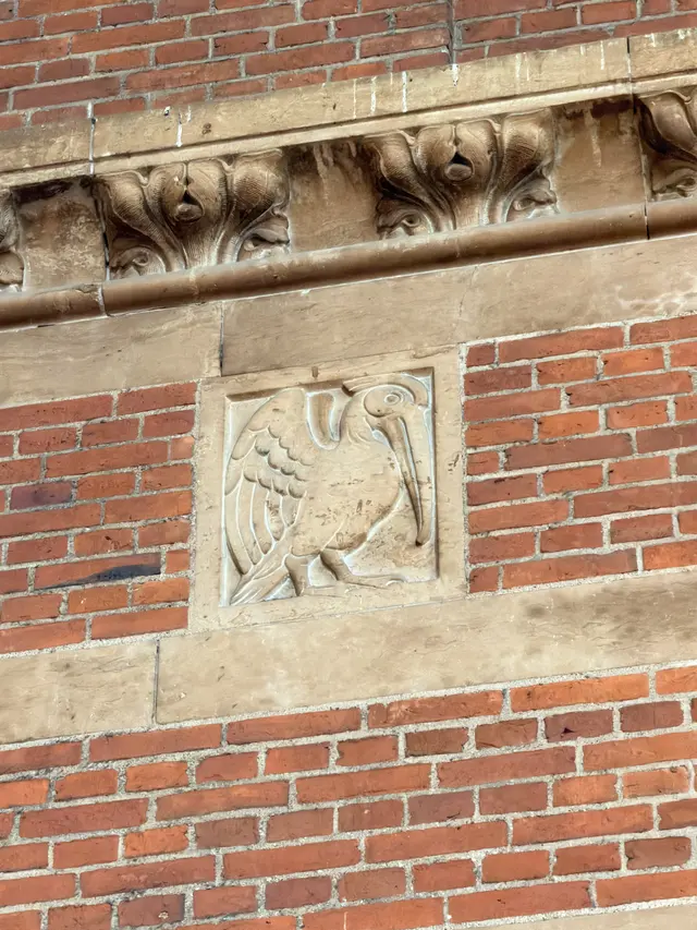 A long-billed wading bird carved in a flat sandstone panel, set flush in the red brickwork.