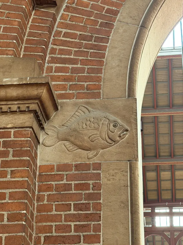 A fish carved in relief in a sandstone block at the springing of a brick and stone arch, with red-painted timber and roof boarding beyond.