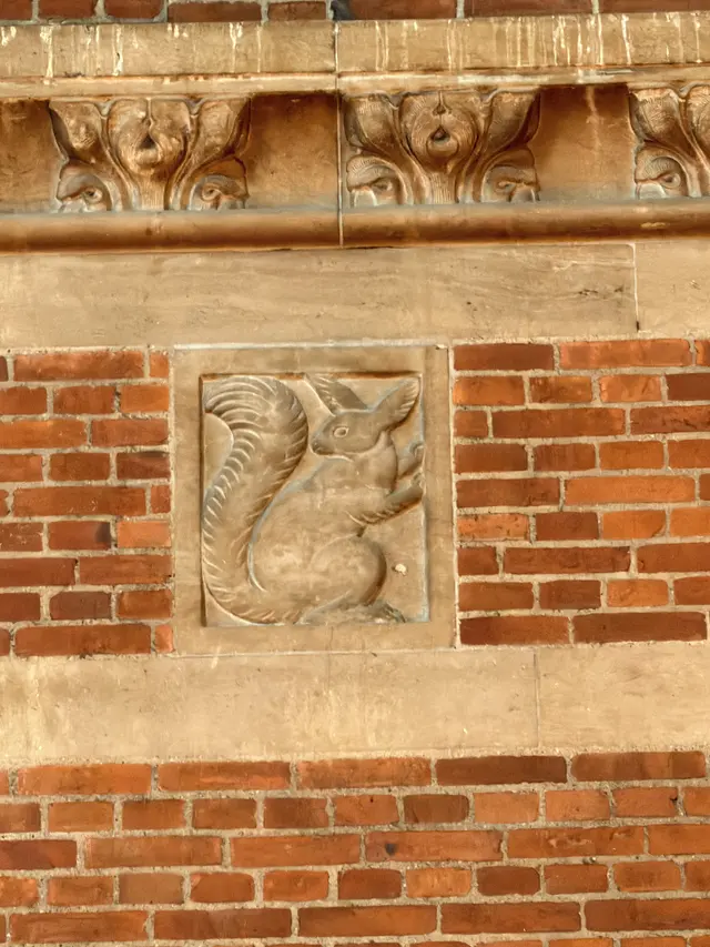 A squirrel with a curled tail carved in a flat sandstone panel, set flush in the red brickwork below a leaf frieze.