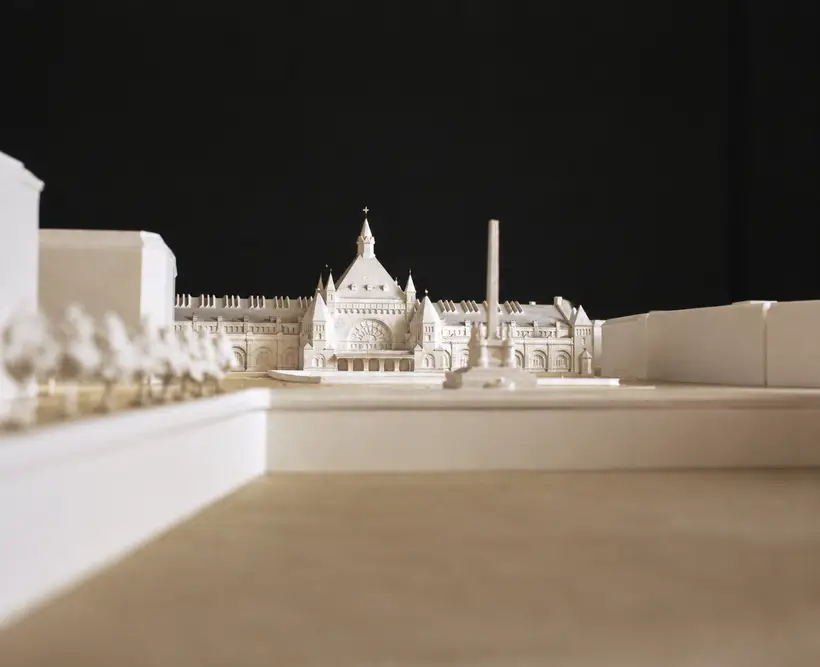 A white architectural model seen from below: a central block with a spire and a round window, low wings, and an obelisk in front.