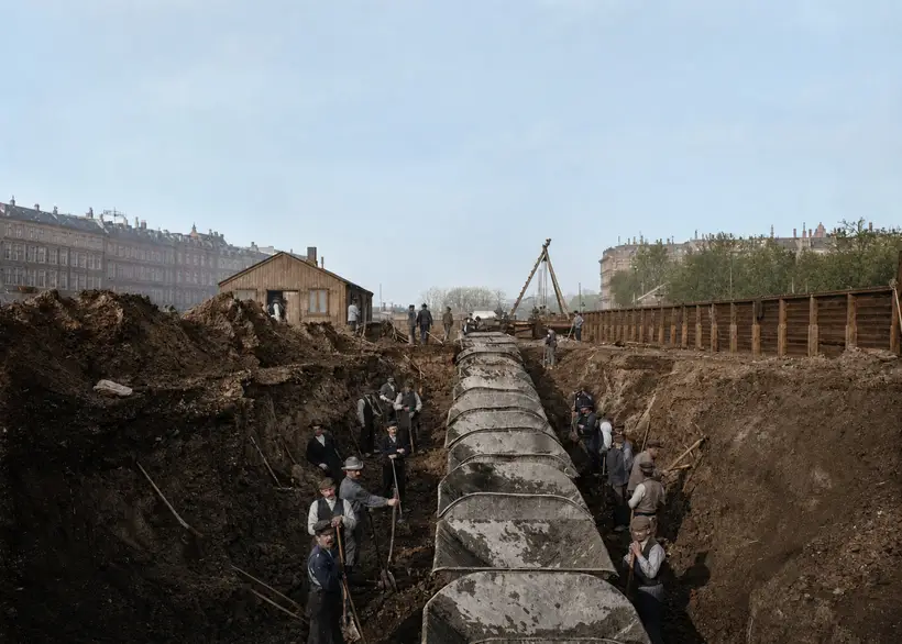Workmen with shovels stand in a deep trench around a line of large pipes.