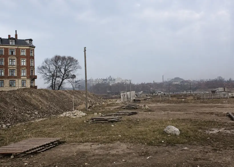 Open cleared ground with earth banks, stacks of timber, and a corner apartment block at left.