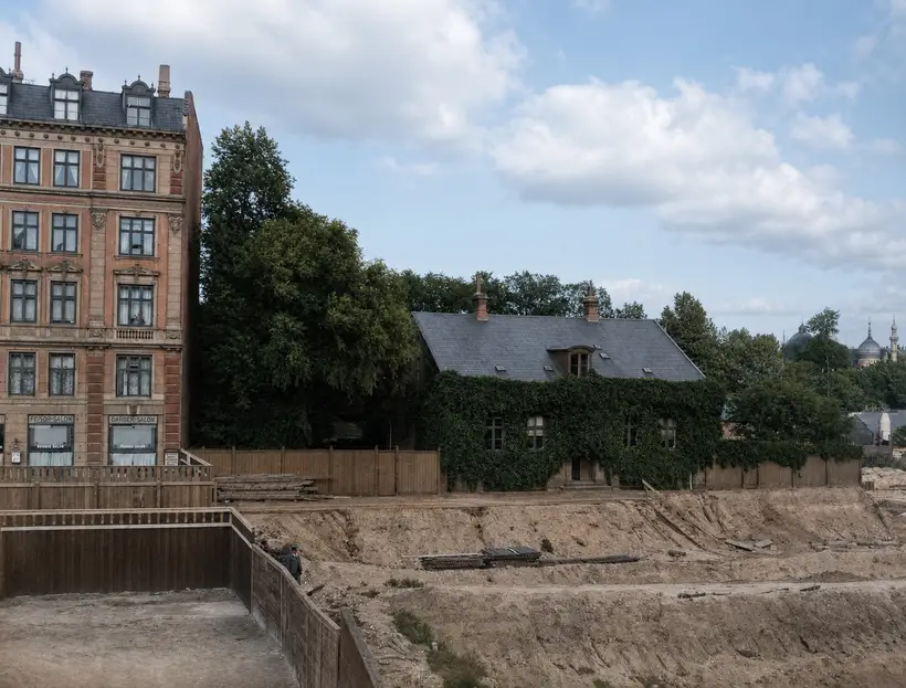Excavated ground along a tall timber hoarding. A five-storey corner block with shop windows at street level stands at left and a low house covered in creeper under a slate roof at centre. Timber and track material lie in the bottom of the cut.