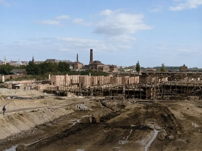 Panorama across a large building site. Long brick walls run the width of the frame with tall round-timber scaffold poles and working platforms at two levels. Churned mud and standing water in front, factory chimneys and a domed hall on the skyline.