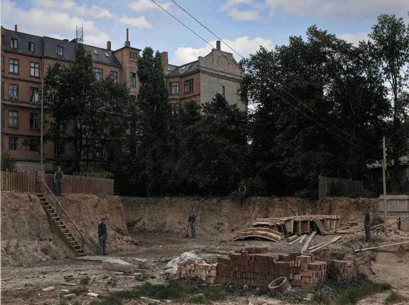 A deep excavation with five workmen. A long timber stair climbs the bank to a picket fence. Stacks of brick and timber, some of it curved, lie at right. Red brick apartment blocks and tall trees stand beyond the fence.