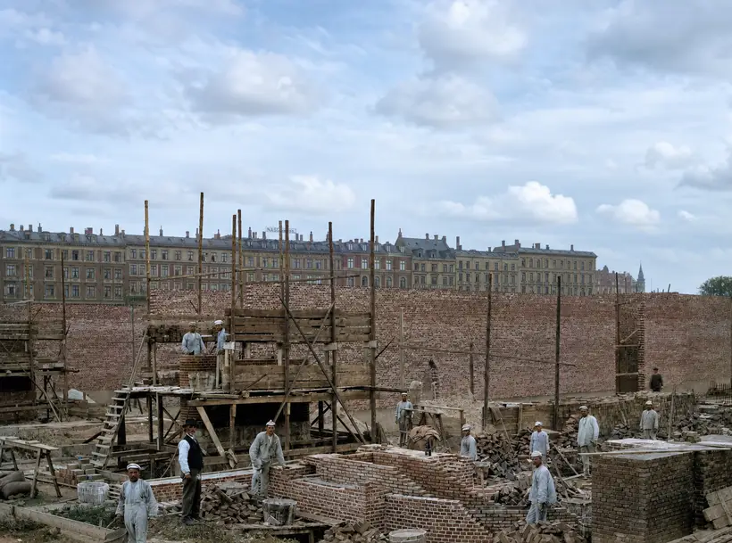 A dozen or so masons in pale working clothes laying brick on low walls and on a raised timber platform. Tall round-timber scaffold poles and a long brick wall behind, with a row of apartment blocks above.