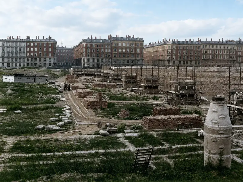 A broad grassed building site with scattered cut stone and large stacks of brick. A horse and cart move along a dirt route. A long wall with brick piers and timber scaffolding stands at right, with a row of apartment blocks and a street opening beyond.