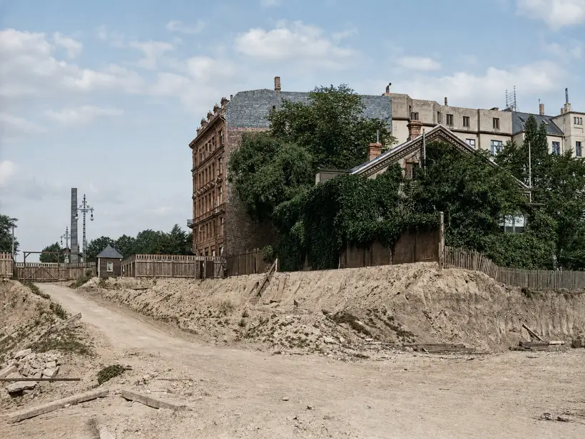 A broad unmade works road at the bottom of cut ground. A raw earth slope rises at right to a plank fence, with a tall dark apartment block and a creeper-covered house behind it. A gate, a small hut and a lattice mast stand at left.