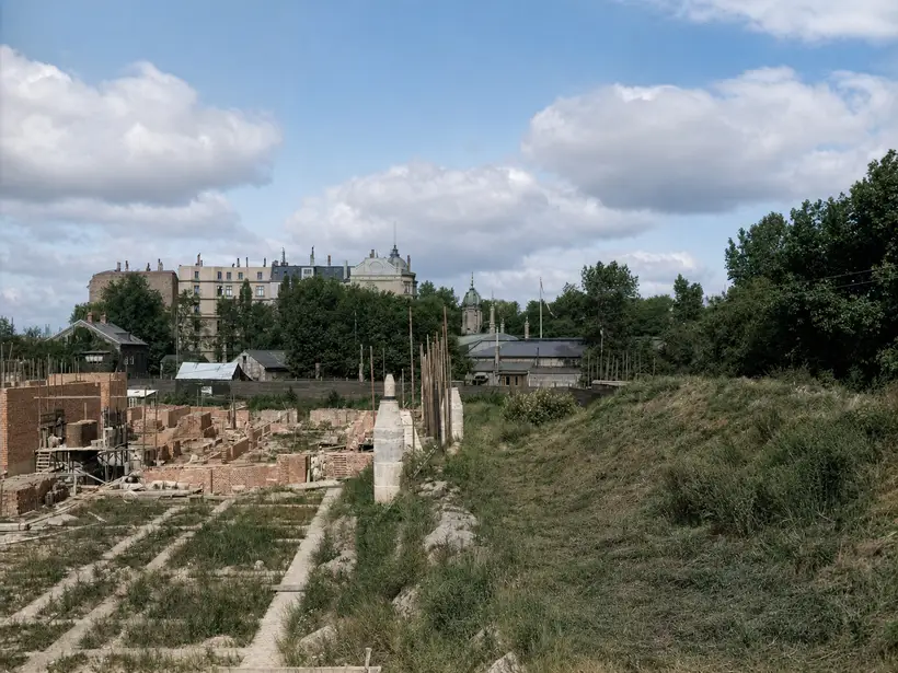 A building site seen lengthways. Low brick walls and foundations under construction at left, a grassed bank at right. A pale conical upright about the height of a man stands at centre, with apartment blocks, a dome and a green spire beyond.