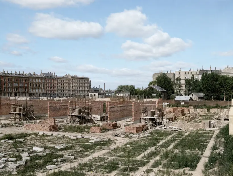 Long walls divided by brick piers, with small timber scaffolds at intervals and brick stacked along the wall foot. A grassed area ruled by pale foundation strips lies in front, with a row of apartment blocks and a factory chimney behind.