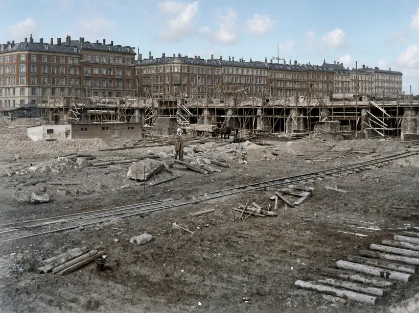 A wide building site: a long line of heavy piers with arched openings behind dense round-pole scaffolding. A horse cart loaded with boards, timber site huts at left, works track and scattered timber across the foreground.