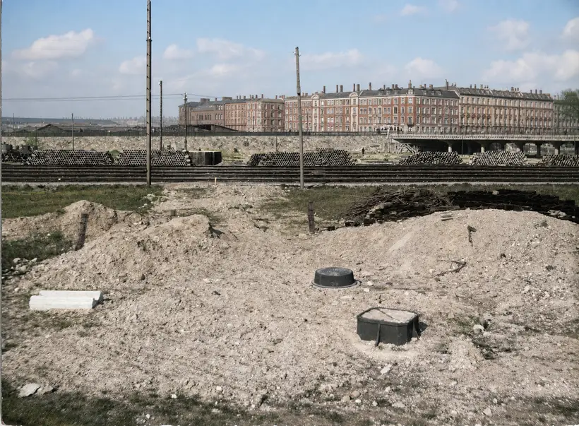 Excavated and back-filled ground with two dark chamber covers in front, railway tracks beyond, long banks of stacked pipes, telegraph poles, a low road bridge at right and a terrace of apartment blocks on the horizon.