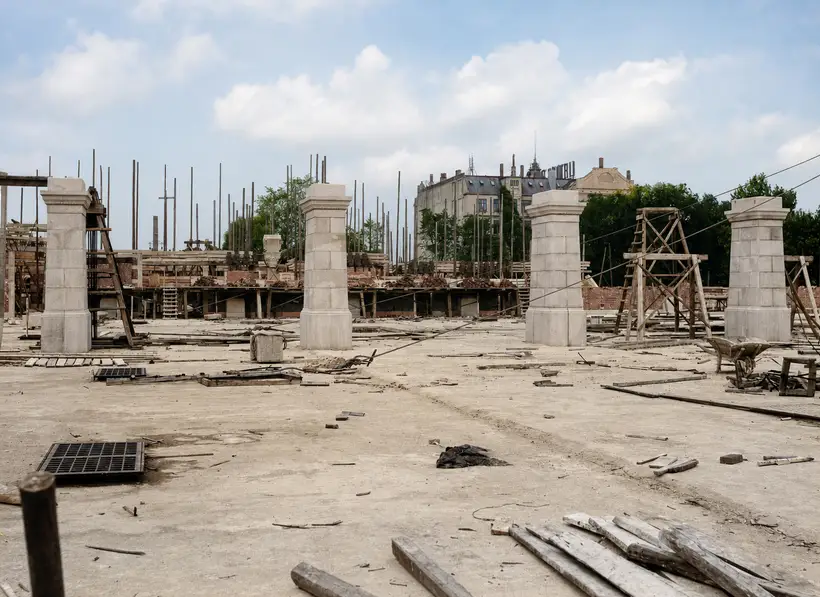 Four pale stone piers stand free on a broad deck. Behind them a low brick wall and a dense forest of scaffold poles; at right a braced timber hoisting frame and a wheelbarrow.