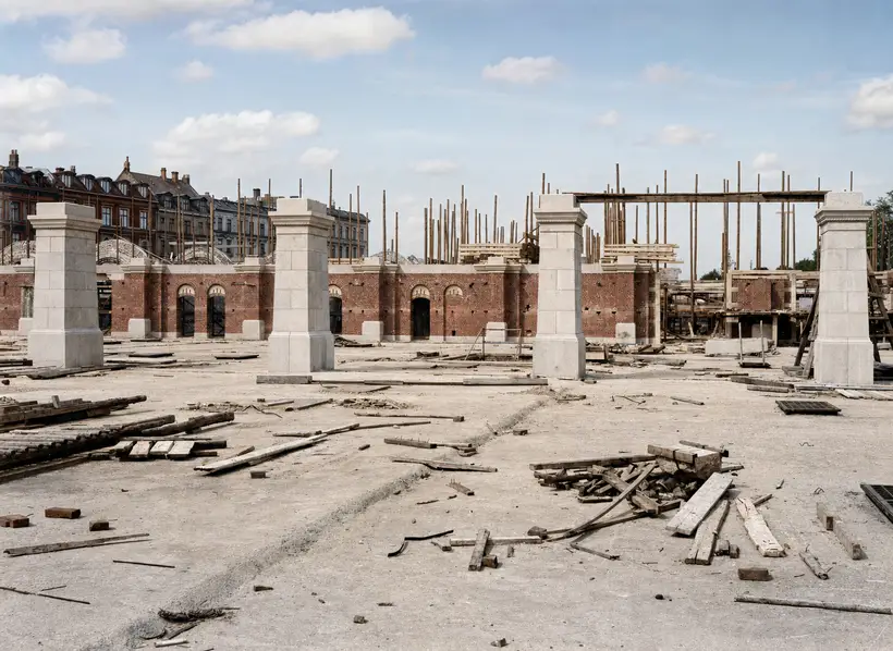 Four stone piers on an open deck in front of a long arched brick wall with a heavy stone coping. Close-set scaffold poles and a large timber portal frame behind, heaps of timber and boards in front.